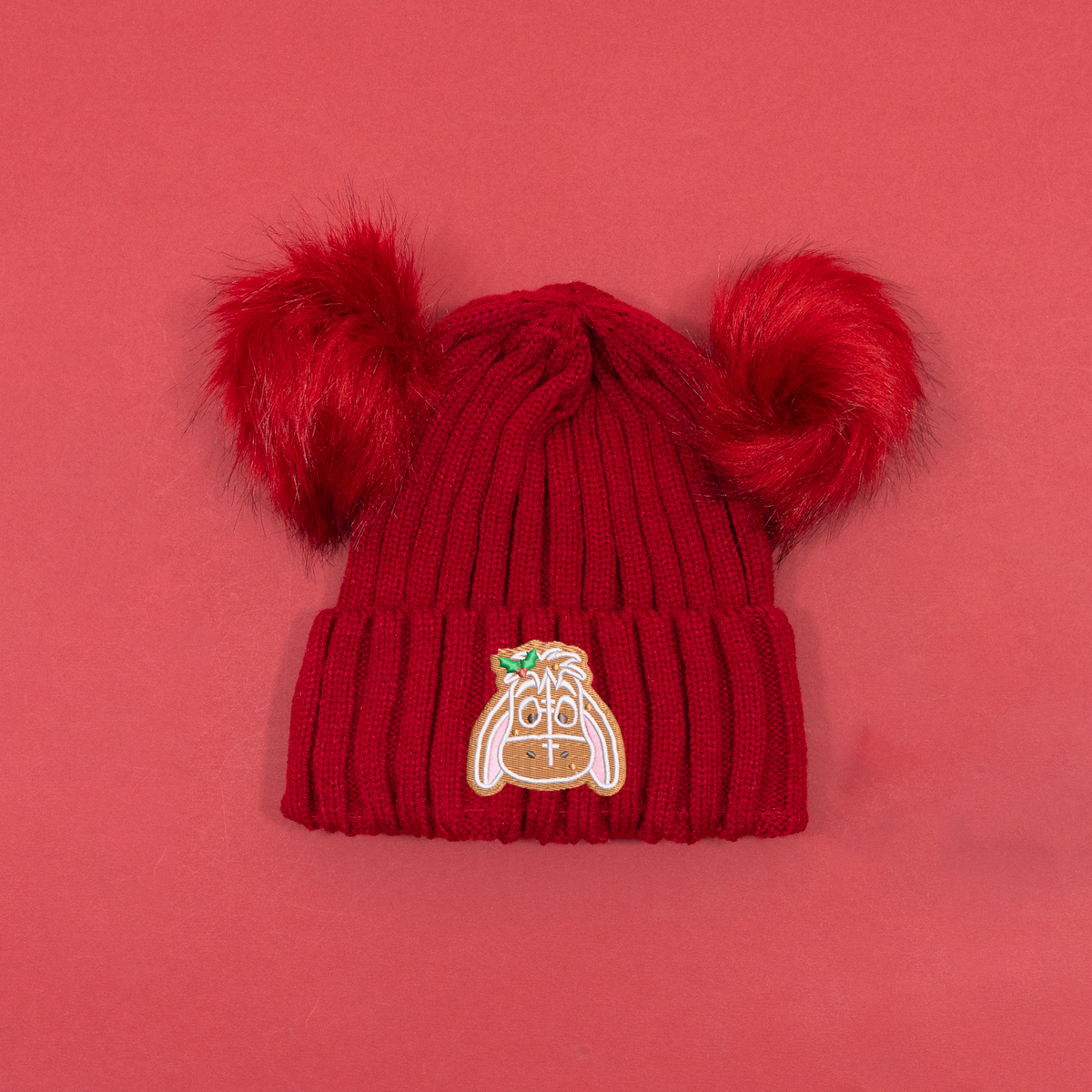 Red ribbed knitted beanie with two fluffy red pom poms and Eeyore gingerbread embroidery with holly on the cuff.