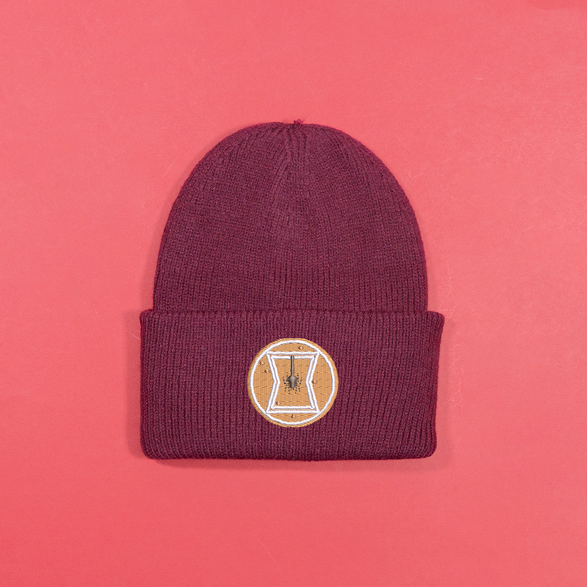 Burgundy knitted beanie with gingerbread-style Black Widow emblem embroidery on the turned-up cuff.
