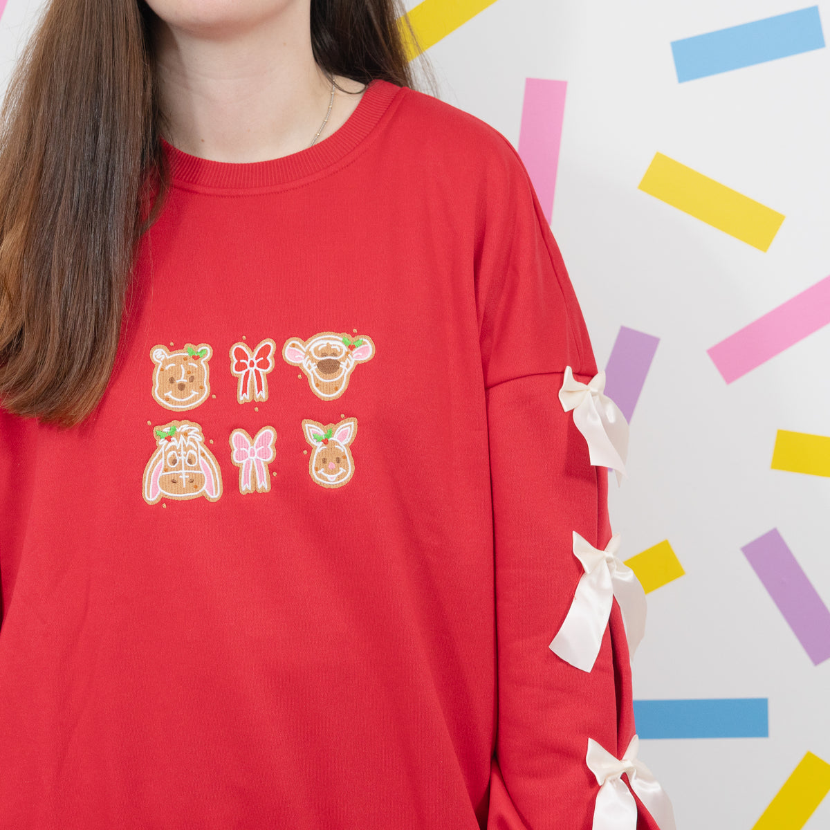 Modelled close-up of the red Winnie and Pals jumper showing gingerbread character embroidery and white sleeve bows.