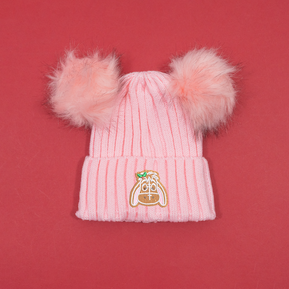 Pink ribbed knitted beanie with two fluffy pink pom poms and Eeyore gingerbread embroidery with holly on the cuff.