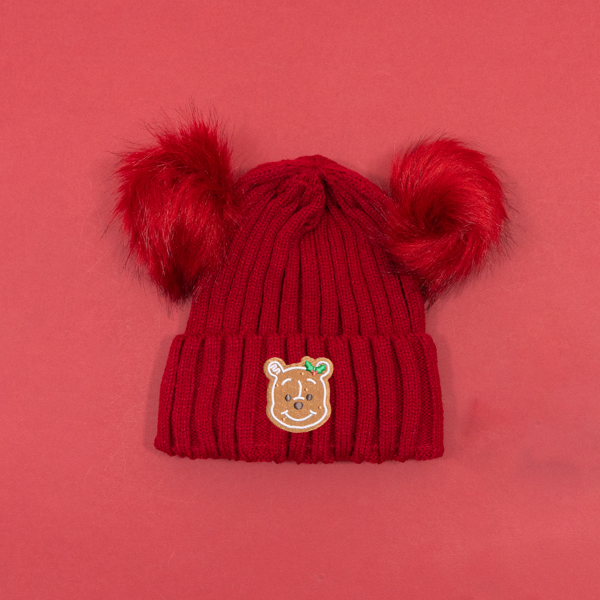 Red ribbed knitted beanie with two fluffy red pom poms and Winnie the Pooh gingerbread embroidery with holly on the cuff.